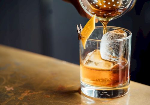 A whiskey tumbler with ice and an orange twist, as a bartender pours amber liquid from a shaker, on a rustic wooden bar.