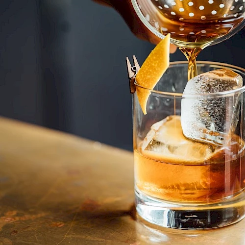 A whiskey tumbler with ice and an orange twist, as a bartender pours amber liquid from a shaker, on a rustic wooden bar.