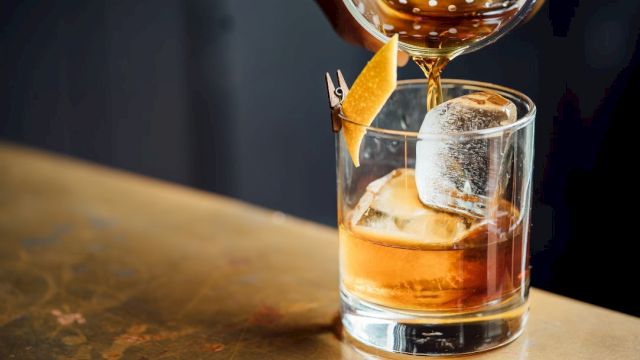 A whiskey tumbler with ice and an orange twist, as a bartender pours amber liquid from a shaker, on a rustic wooden bar.