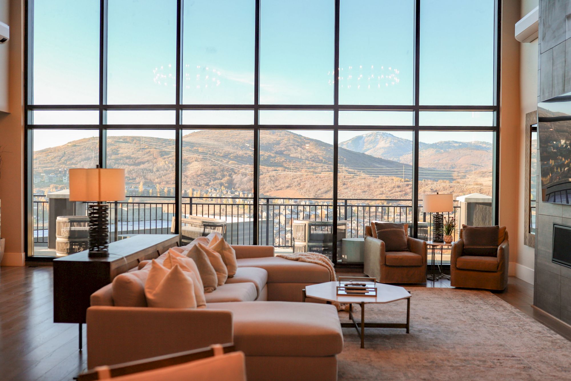 A spacious modern living room with large floor-to-ceiling windows, comfy beige sofas, a coffee table, and views of rolling hills outside.
