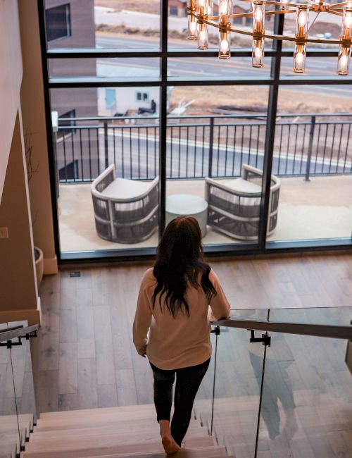 A woman walks down glass stairs toward a bright living area with a large window, sofa, dog, and a balcony view downstairs.