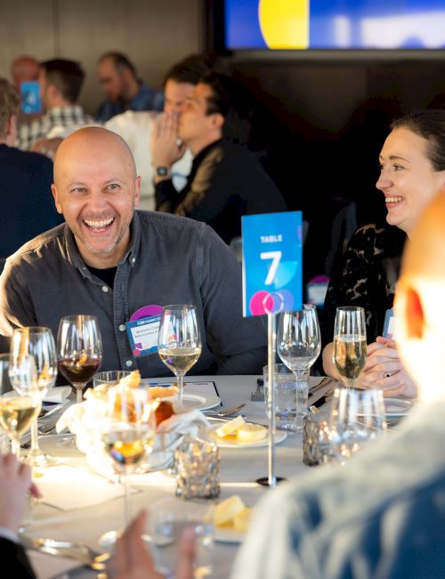 A group of people at a formal dinner laugh and chat around a table with wine glasses; table number 7 sign stands in the center.