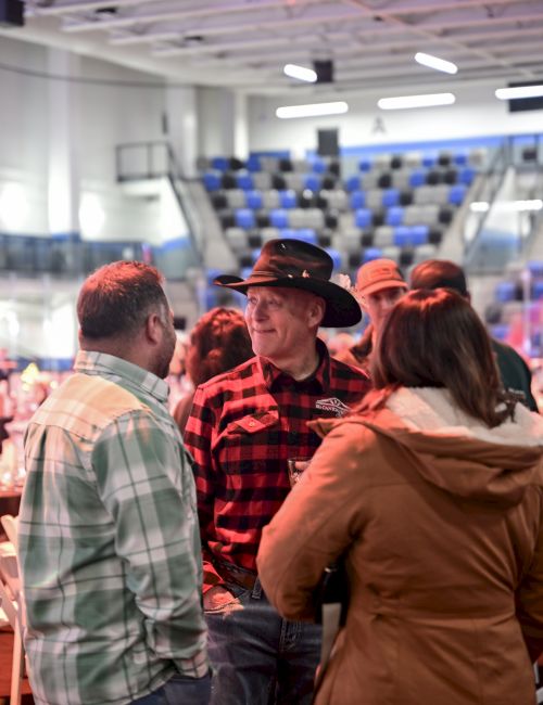 A group of people chat in a crowded indoor event space; one man wears a black hat and red plaid shirt, others in jackets surround him.