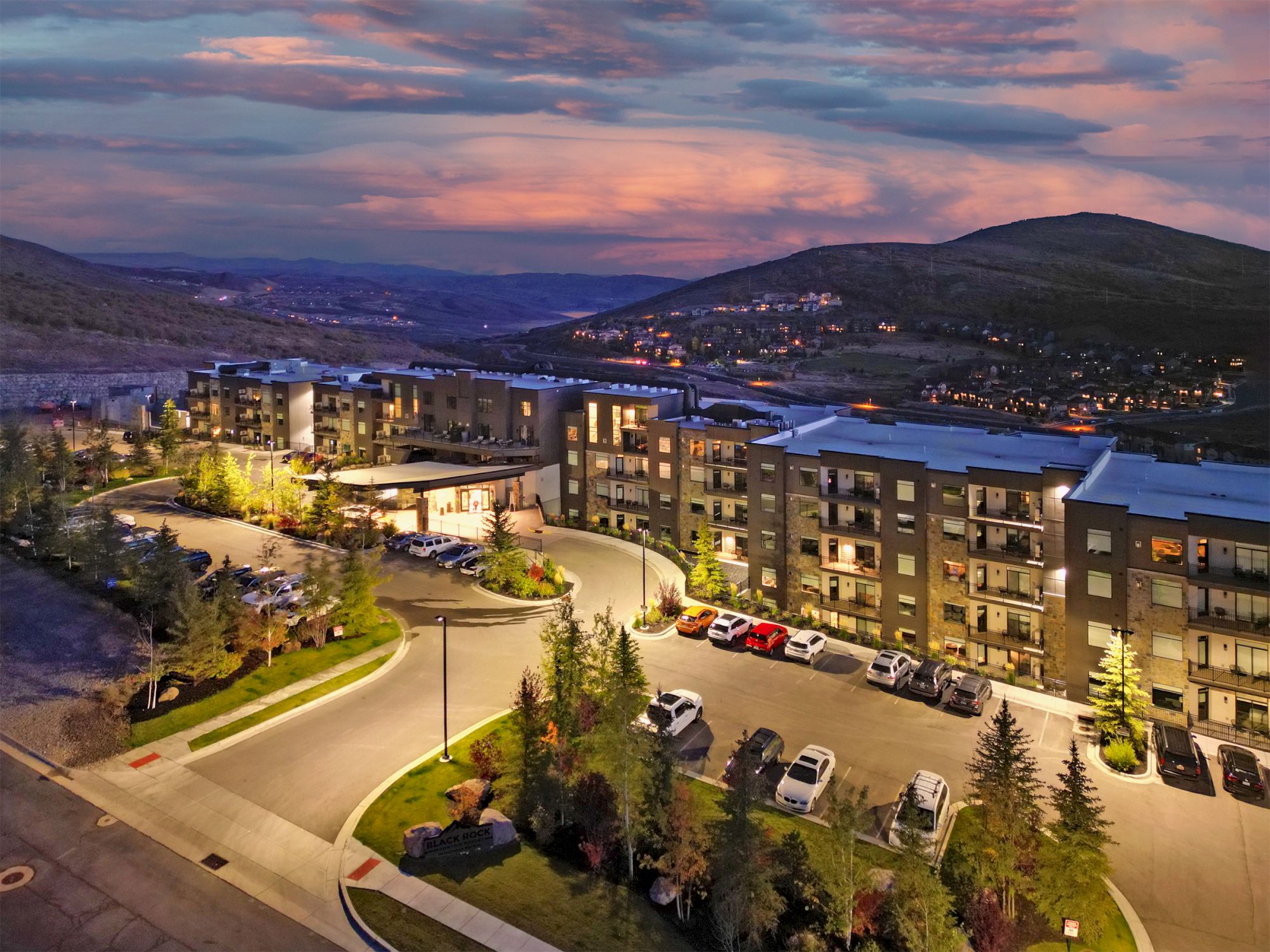 Aerial view of a modern apartment complex at dusk, with lit windows, a curved driveway, parked cars, trees, and a hill in the background.