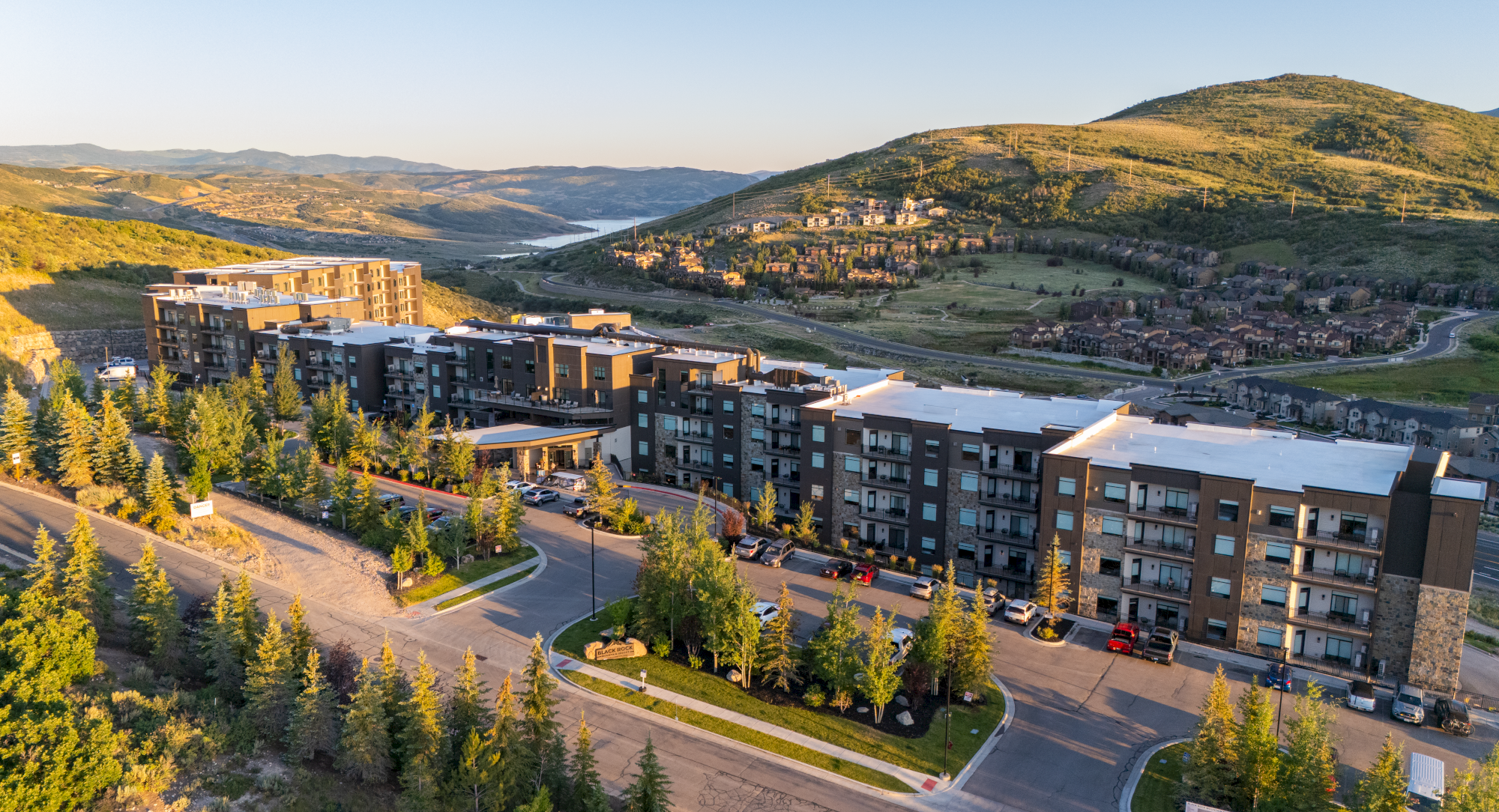 A modern apartment complex sits along a curving road, surrounded by trees, rolling hills, and a distant river under a clear blue sky.