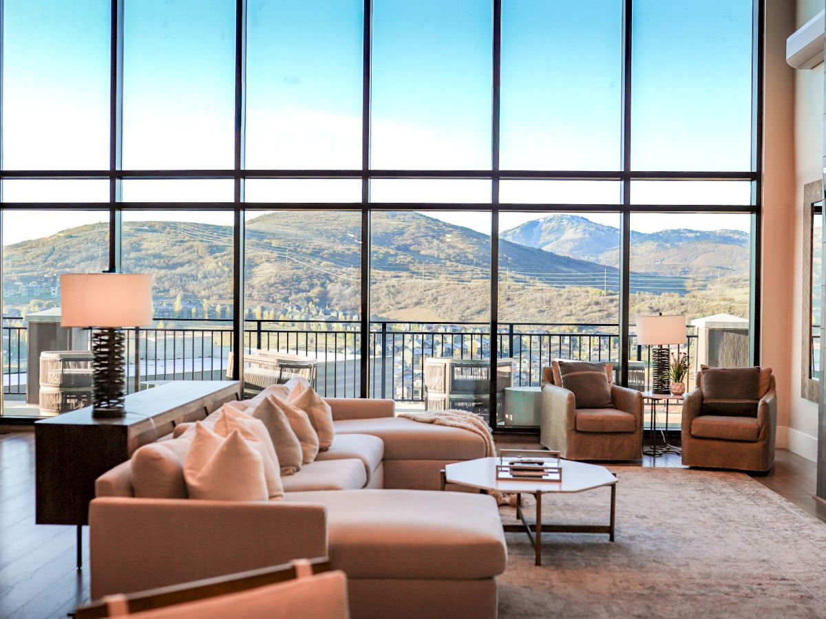 A bright, modern living room with expansive floor-to-ceiling windows, overlooking hills; beige sofas, a coffee table, and stylish lamps.