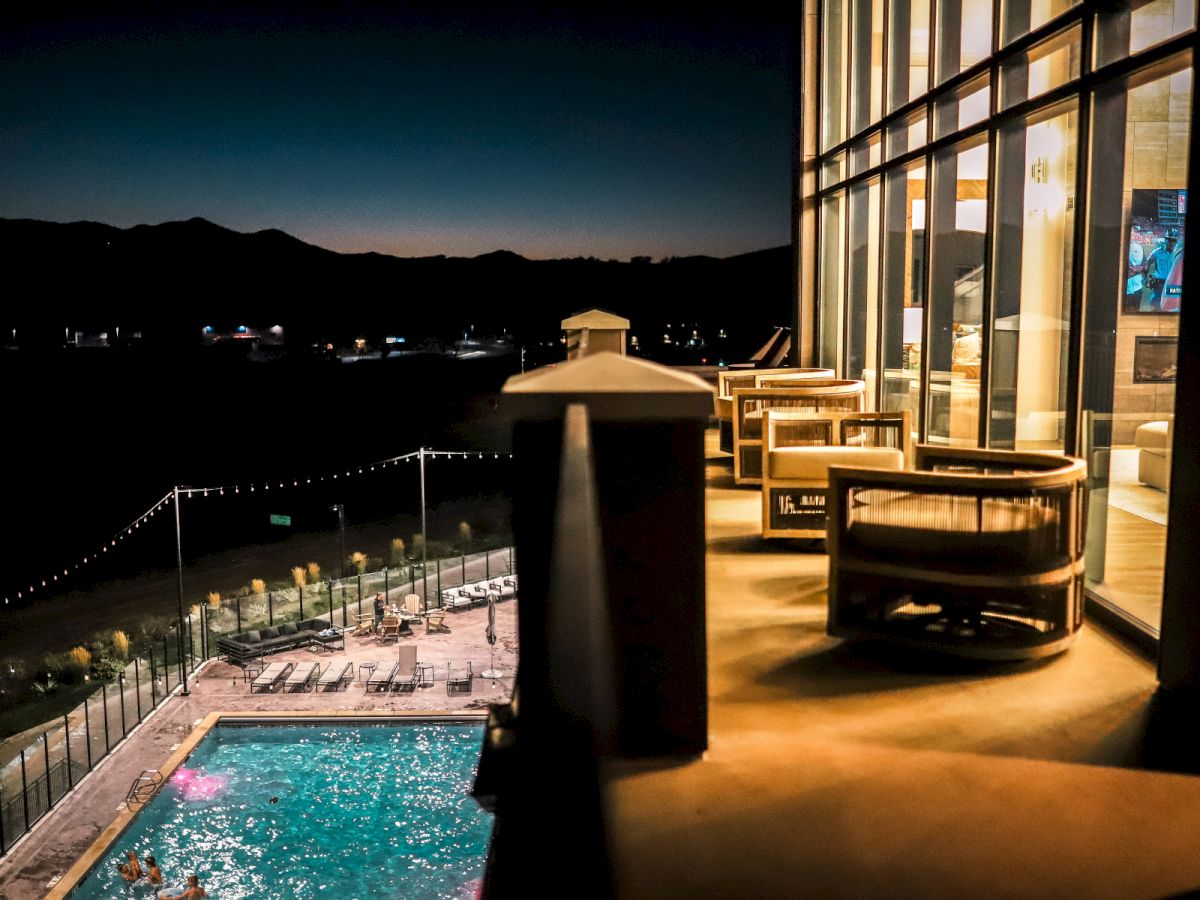 A nighttime hotel balcony overlooks a lit pool area with lounge chairs, glass railing, and bright indoor seating reflecting warm light.