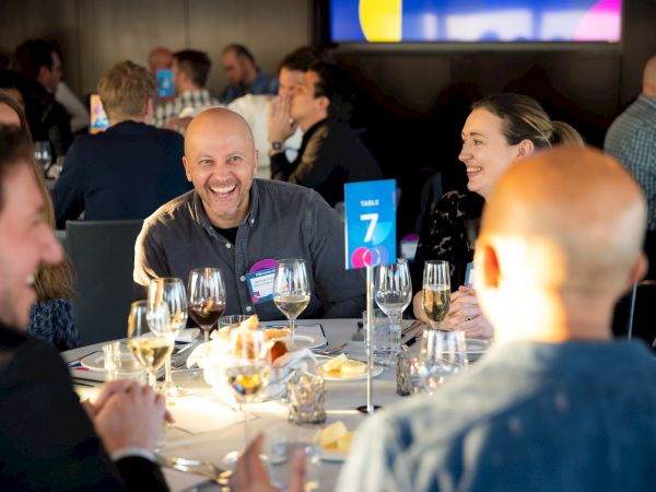 A group of people laughing and chatting around a table at a formal dinner, with wine glasses and a blue table number card, lively atmosphere.