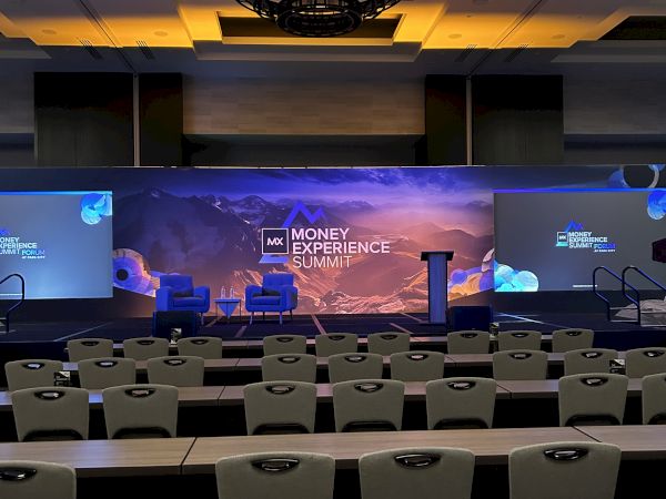 A conference stage with a large screen reading &ldquo;Money Momentum Summit,&rdquo; empty chairs and rows in a dim, auditorium setup.
