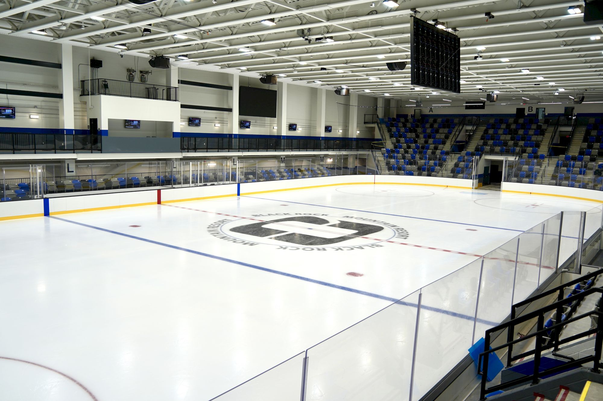 An empty indoor ice hockey rink with seating, boards, goal nets, and a central logo on the ice.