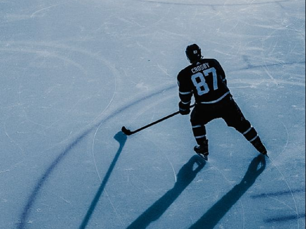 An ice hockey player in black jersey number 87 glides on the rink with a puck, casting long shadows on the blue ice. end.