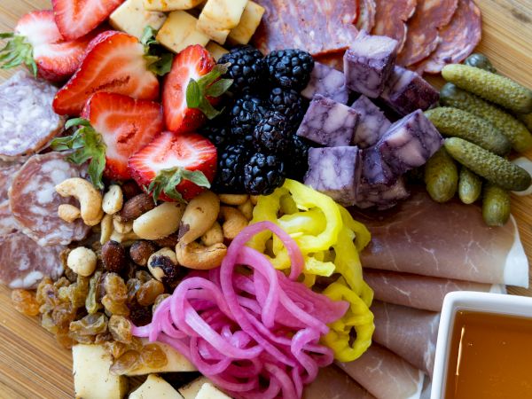 A round charcuterie board with strawberries, cubed cheese, blackberries, pickles, pickled onions, assorted meats, crackers, nuts, and a honey dip cup.