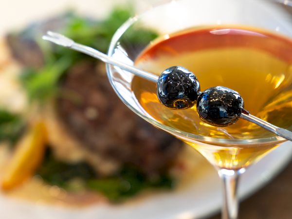 A cocktail with two blackberries on a skewer, in a coupe glass, with a blurred plate of food in the background.