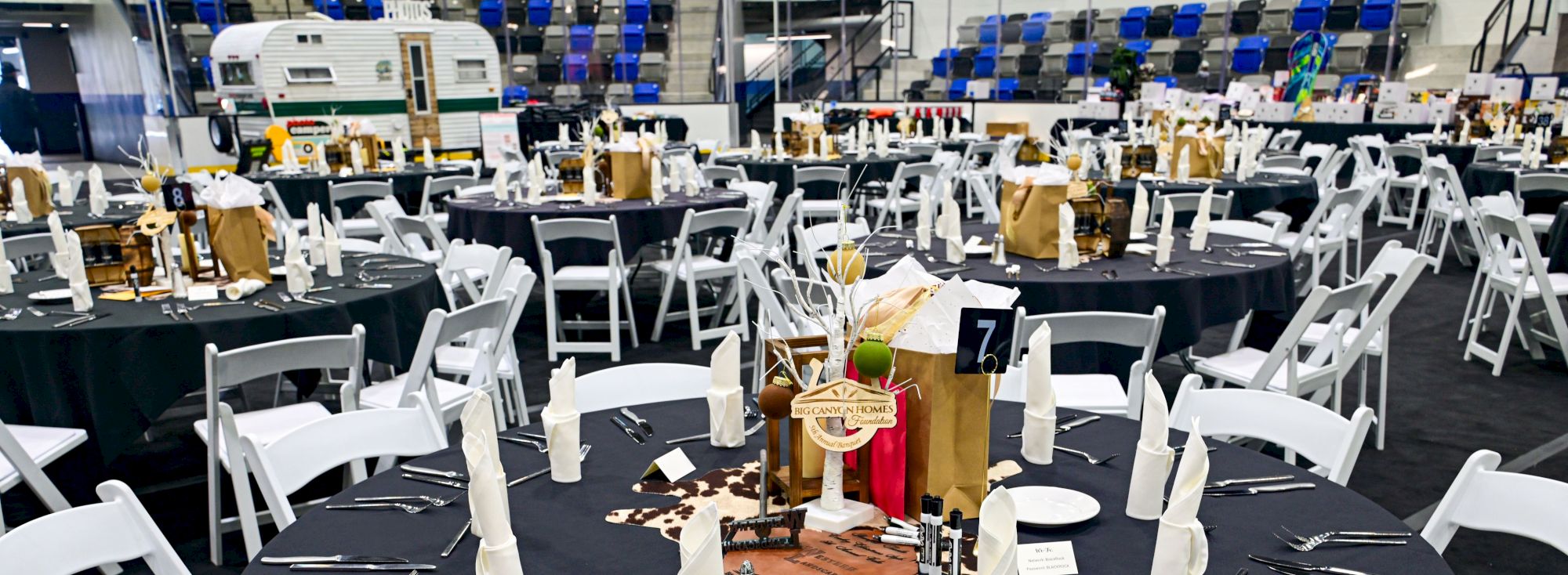 A banquet setup with round tables, black tablecloths, white folding chairs, neatly arranged silverware, napkins folded at each place, and centerpieces with gold accents.