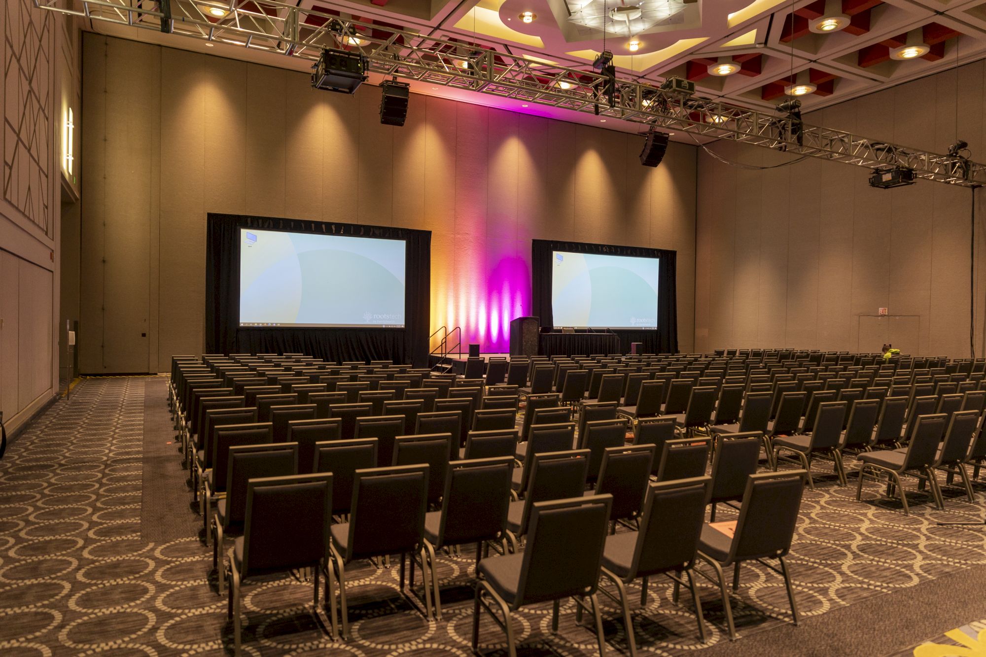 A conference hall with rows of black chairs facing two large screens, stage lighting in purple hues, and patterned carpet, empty seats, and projector setup.