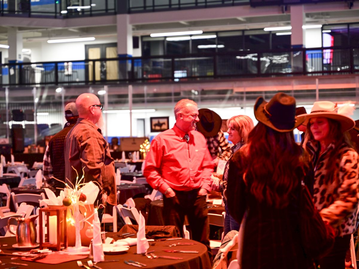 People mingling at a social event in a large hall with round tables, decorations, and guests chatting under bright lights.