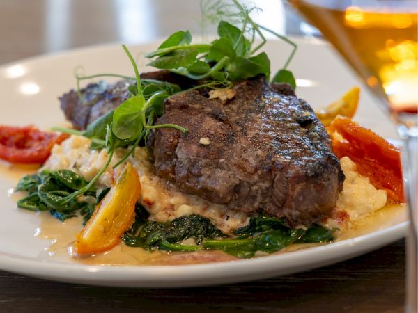 A plated steak sits atop mashed potatoes with greens, surrounded by colorful vegetables, on a light table with a glass of wine in the background. This is a delicious-looking dish.