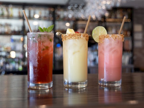 Three colorful cocktails on a bar, each with a rim garnish and a straw, rowed against a blurred back bar scene.