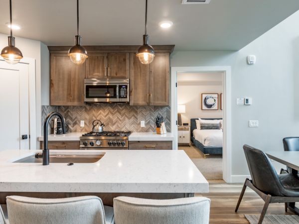 A modern kitchen with a white island, black faucet, stainless appliances, wooden cabinets, pendant lights, and a view into a bedroom.