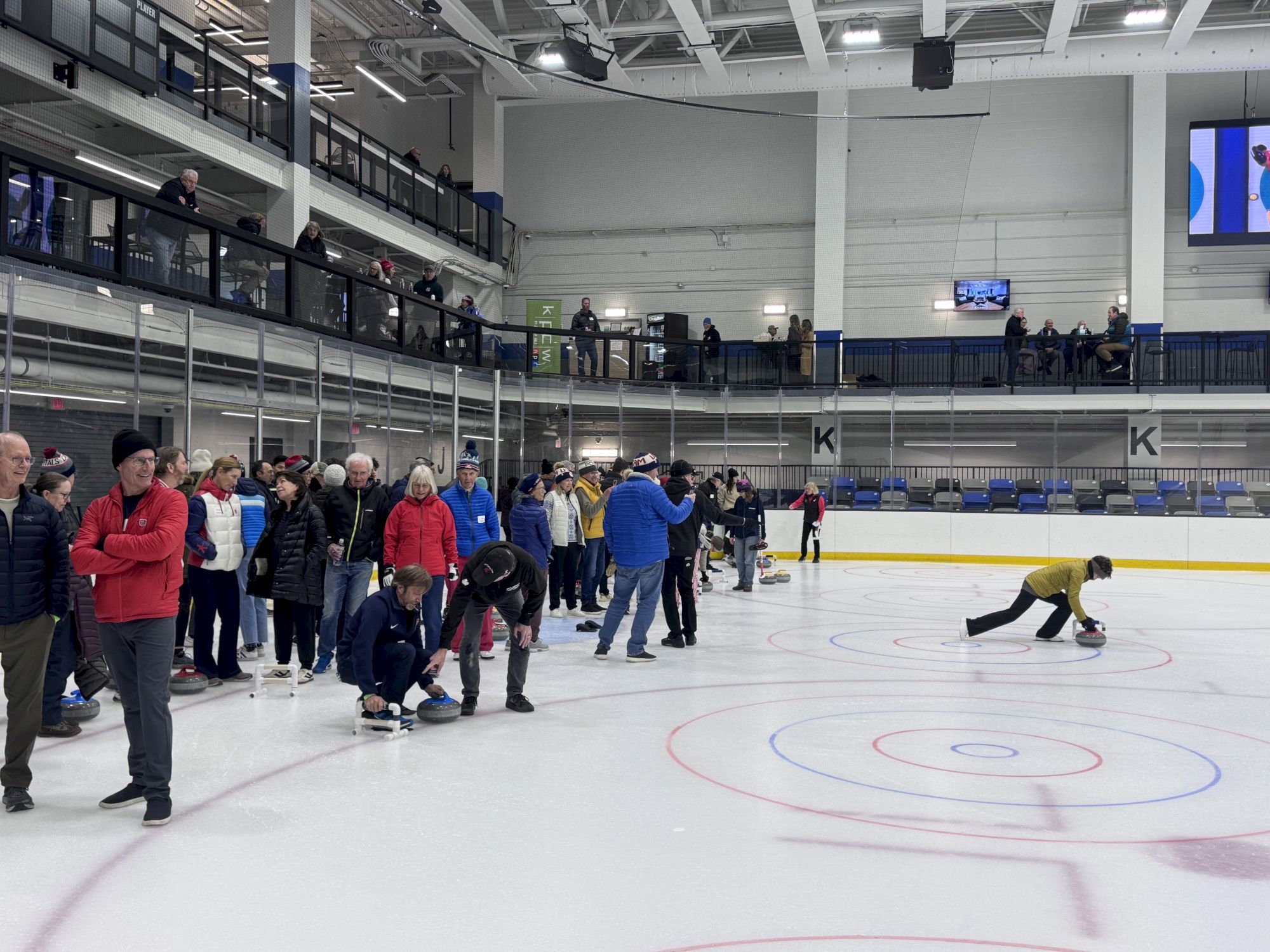 A line of people on an ice rink watch a skater in a yellow suit sweeping the ice, while others chat and queue along the boards.