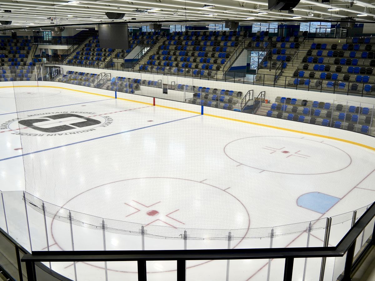 An empty indoor ice hockey rink with protective glass, blue seats, and yellow dasher boards around the ice, ready for a game.