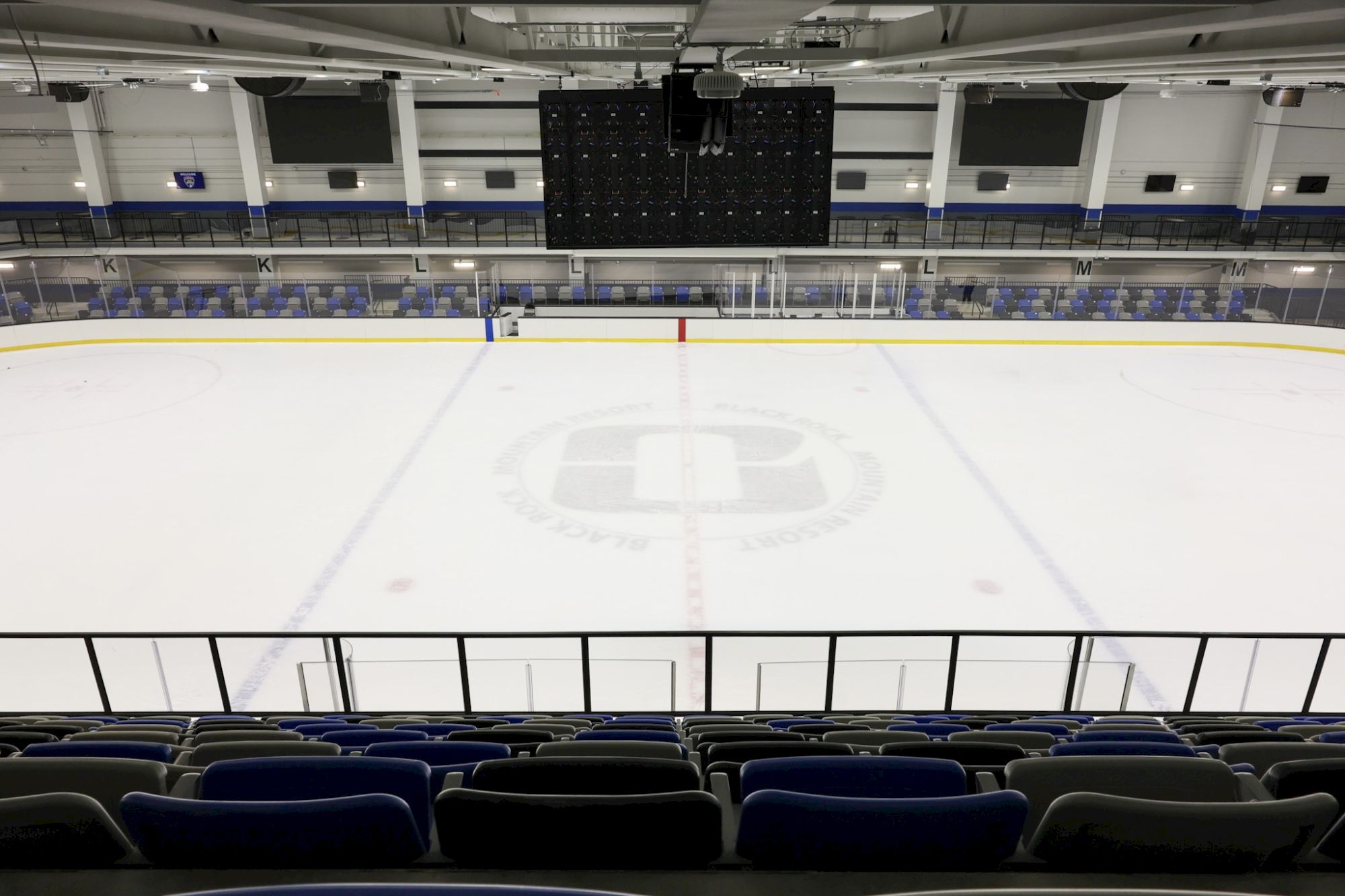 A pristine indoor ice hockey rink with empty blue seats, boards, and a large ceiling scoreboard above the ice, ready for a game.