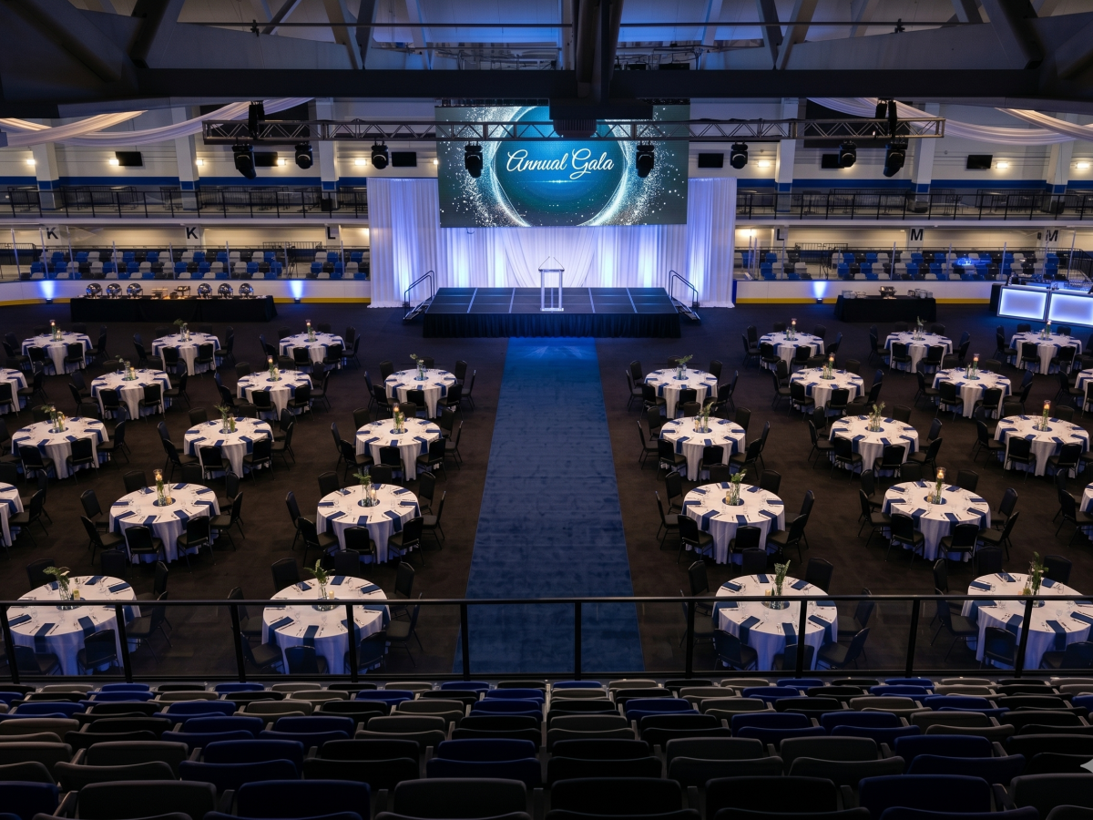 A large banquet hall with round tables, blue accents, a long blue carpet to a stage, and a big screen reading &ldquo;Event Gala.&rdquo;