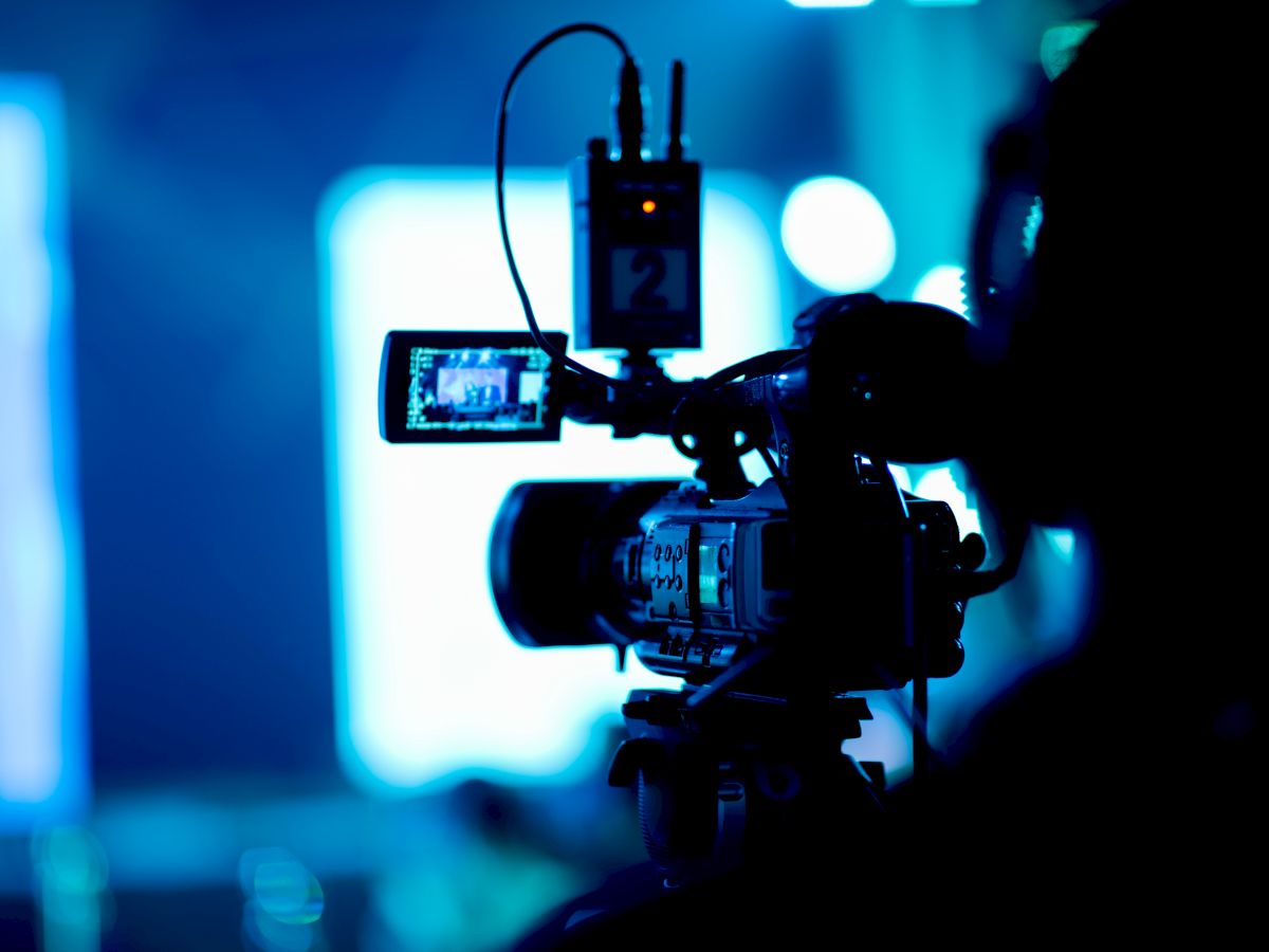 A video camera operator filming on a dimly lit set, monitor and rig visible, blue stage lights in the background, action captured on camera.