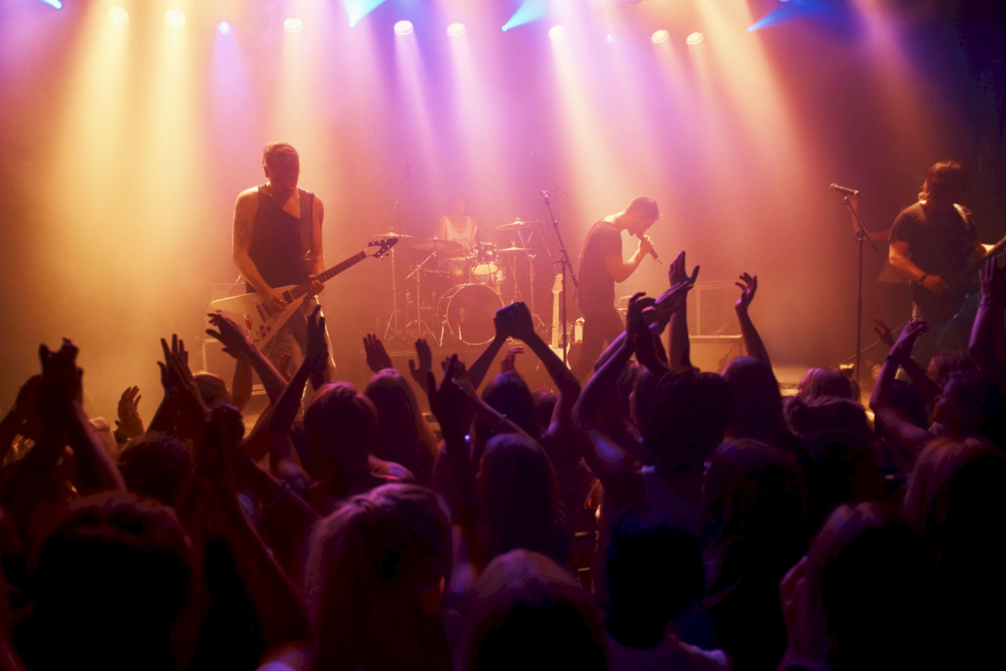 A live rock band performs on stage with colorful lights while a crowd raises their hands and cheers in front of them.