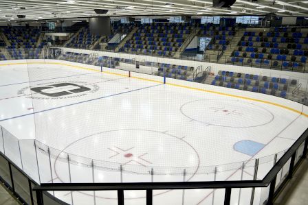 An empty professional ice hockey rink with protective glass, blue seating, and overhead lights, ready for a game.