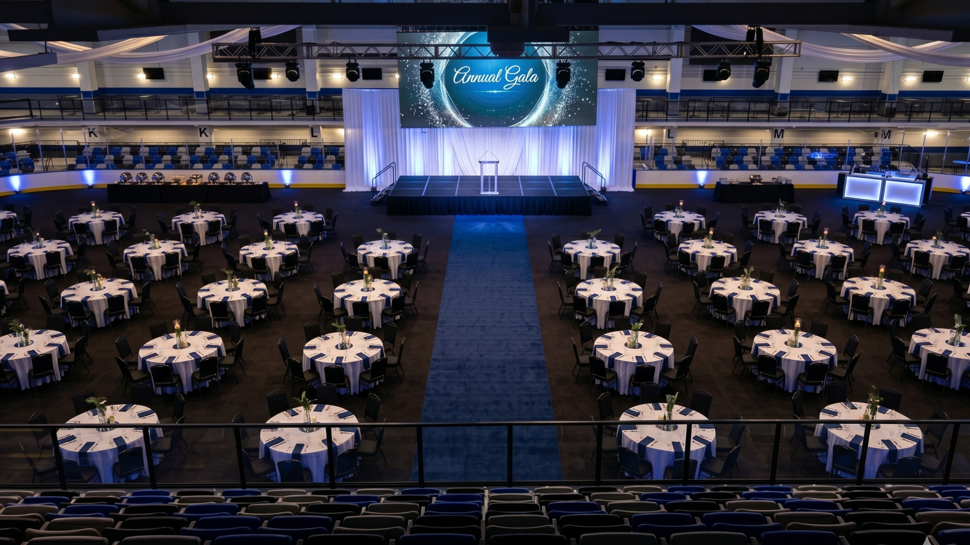 A large banquet hall set for a formal event with round tables, white linens, and blue accents, a stage with a screen top center, and theater seating above.