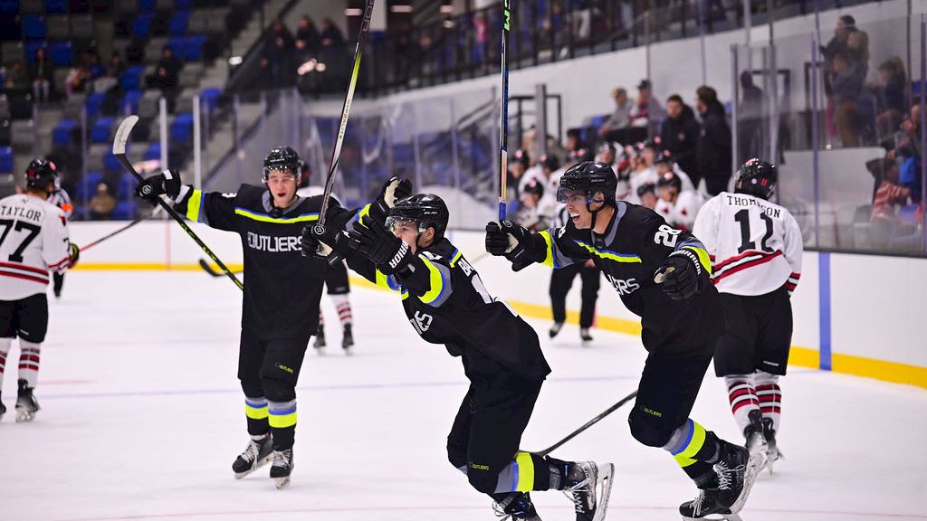 Ice hockey players in dark jerseys celebrate on the rink, raising sticks as they smile in front of the crowd.