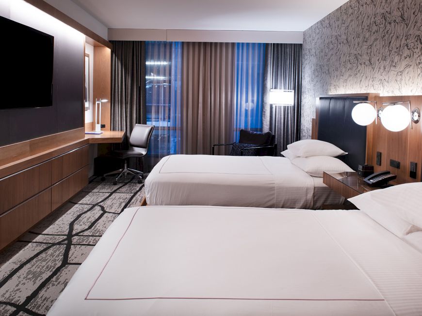 A modern hotel room with two beds, a wall TV, and nightstands lit by lamps and curtained windows.