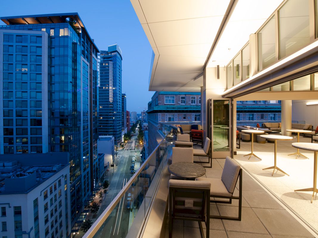 A high-rise balcony overlooks a downtown street and skyscrapers, with modern patio seating and a bright sky at dusk.